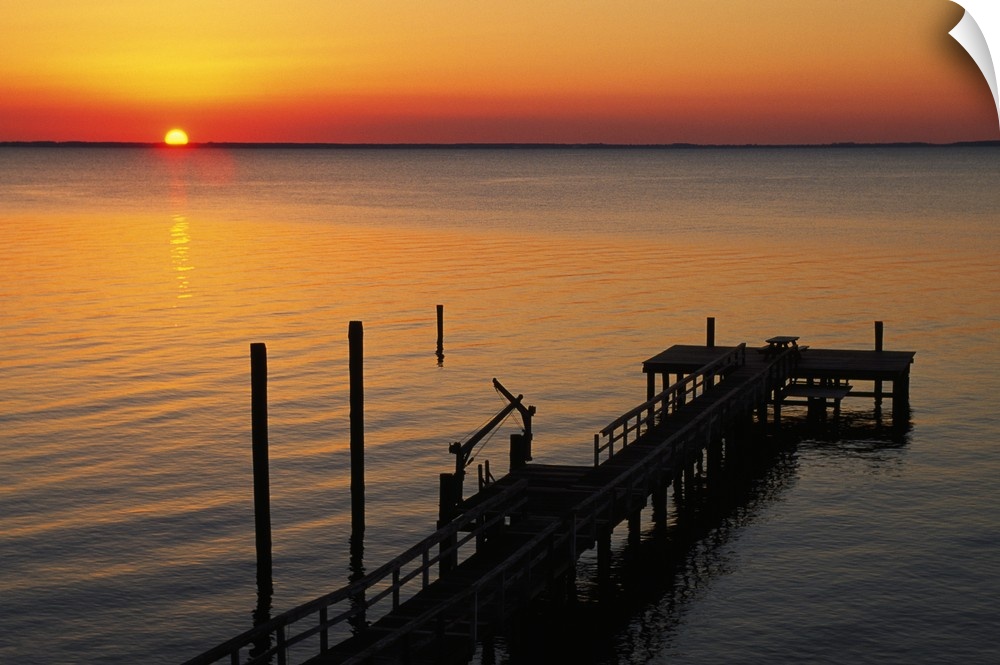 A long pier is photographed reaching out into the ocean with the sun setting just on the horizon.