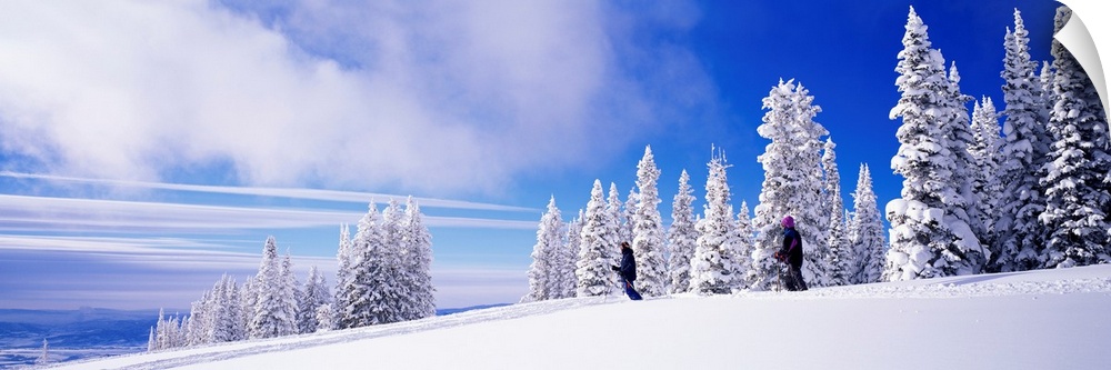 Panoramic photograph focuses on two people skiing on a mountain recently blanketed with fresh snow.  Behind the people is ...