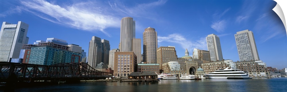 The Boston skyline is photographed in panoramic view from the waterfront during a bright sunny day.