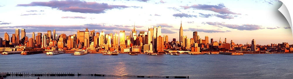Panoramic photograph of the distant Manhattan skyline at the waters edge, with golden skyscrapers as the sun sets.