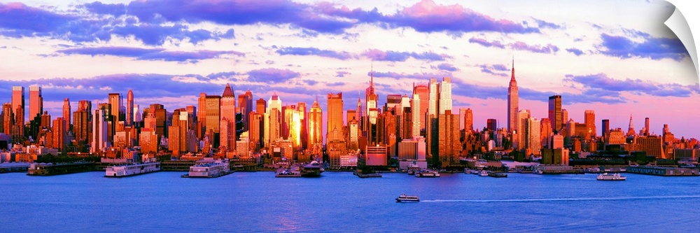 Panoramic photograph of the water in front of the New York City skyline, beneath a vibrant, partly cloudy sky at sunset.