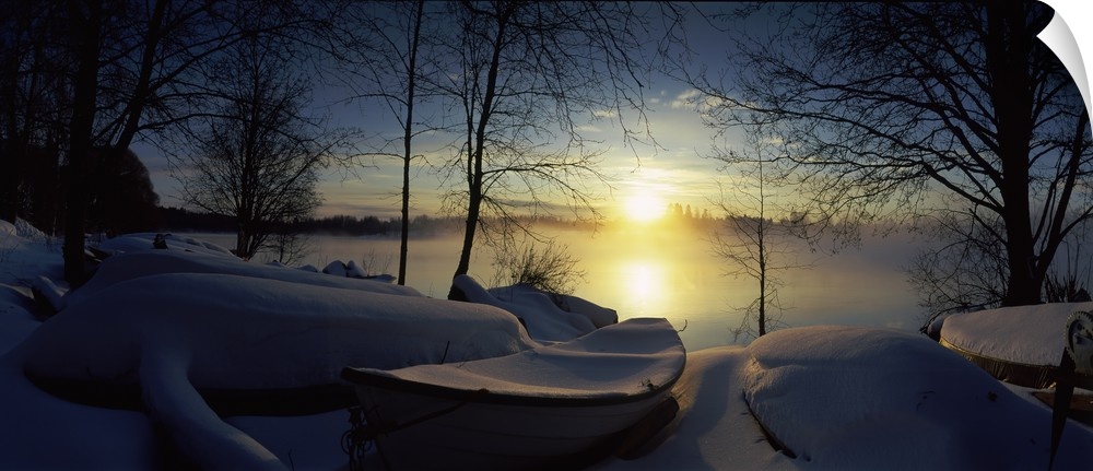 Panoramic photograph displays a group of rowboats blanketed in fresh powder sitting near the shore of a river in Europe.  ...