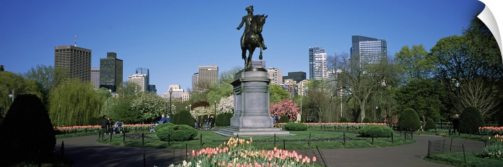 A large statue that stands in the middle of a garden is photographed in panoramic view with tall buildings visible in the ...