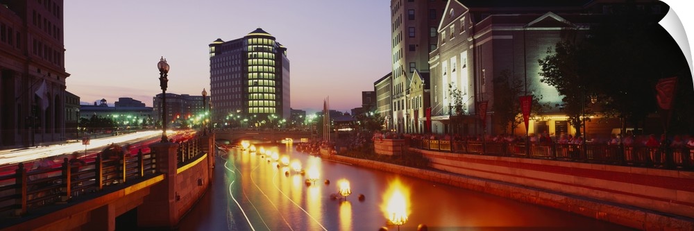 Panoramic canvas photo of lights along a road that leads to buildings in a city at sunset.