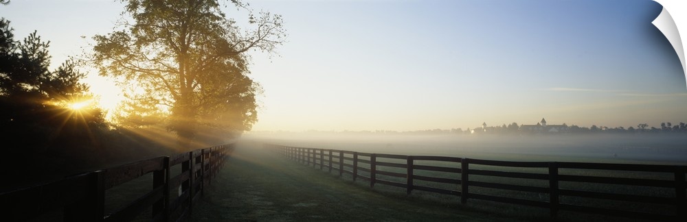 Beams of sunshine reaching across two wooden fences of corrals on a hazy, misty morning in the countryside.