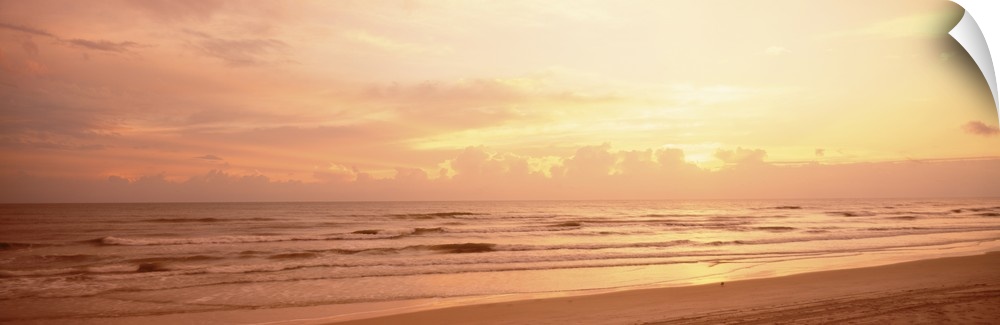 Panoramic photograph of the sun rising above the clouds as small waves break on the sandy shore.