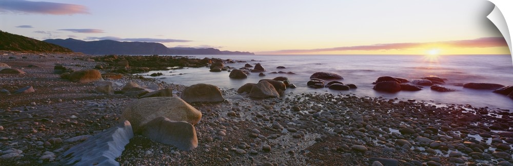 Panoramic photograph of rock and pebble filled shoreline with sun setting in the distance.