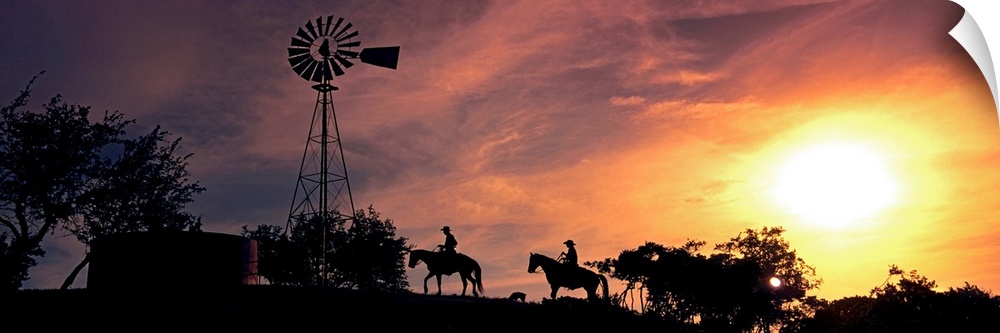This panoramic photograph depicts an evening moment on ranch hill side as horseback riders pass by a windmill trees.