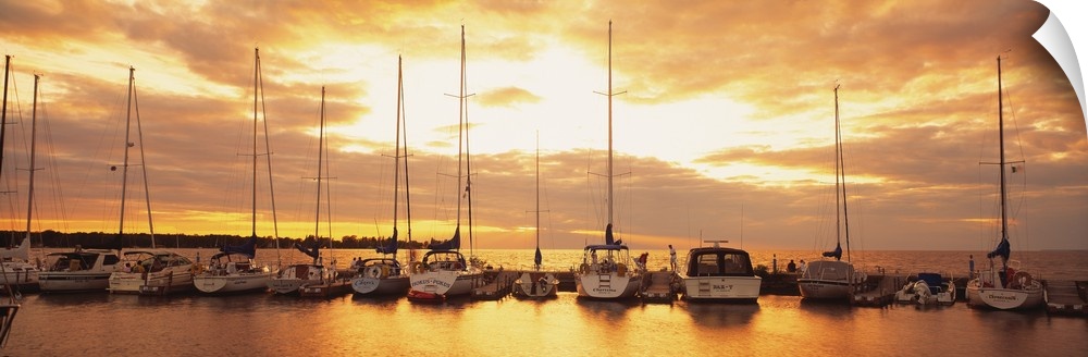 Horizontal panorama of a row of sailboats with their sails down, docked at a port on a lake at sundown in New England.