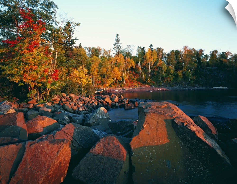 The sun illuminates rust colored rocks and autumn trees at a rocky lake side beach in this landscape photograph.