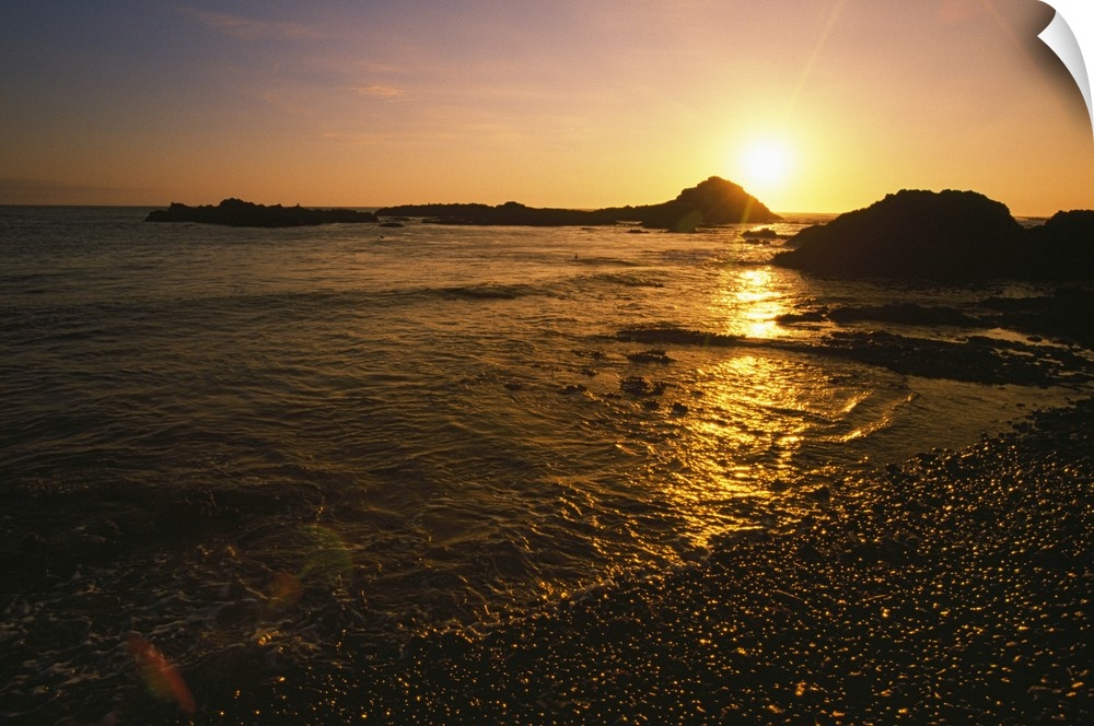 Large, landscape photograph of the sun setting over large rocks in the water, near the shore of the Pacific Coast in Oregon.
