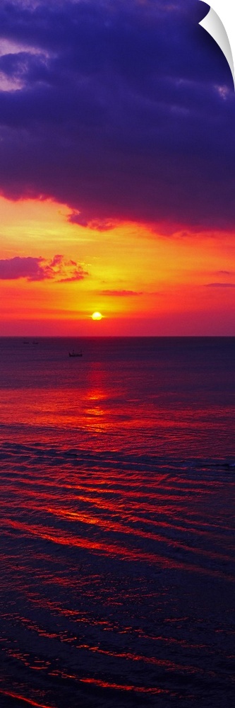 Vertical, large photograph of the sun setting in a vibrant, fiery sky over dark ocean waters, near Bali, Indonesia.