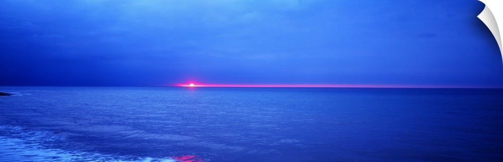 Panoramic photograph of the sun setting just below the horizon over the vast blue ocean.