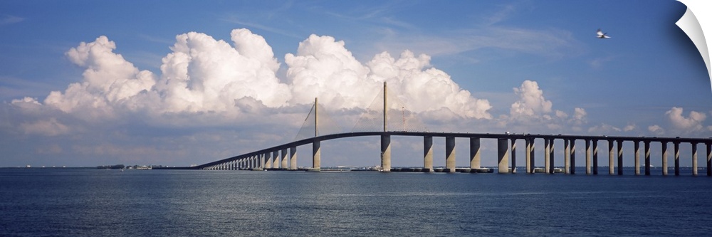 Oversized, horizontal photograph of white fluffy clouds and a blue sky over the Sunshine Skyway Bridge, on the waters of t...