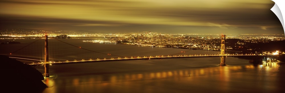 Giant, wide angle photograph of the Golden Gate Bridge lit up at night, the city lights of San Francisco in the background.