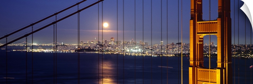 A luminescent full moon shines over the distant city towards the illuminated tower and hanging cables at night in the Bay ...