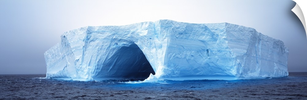 This panoramic photograph is of an immense iceberg that has a tunnel going through the middle of it.