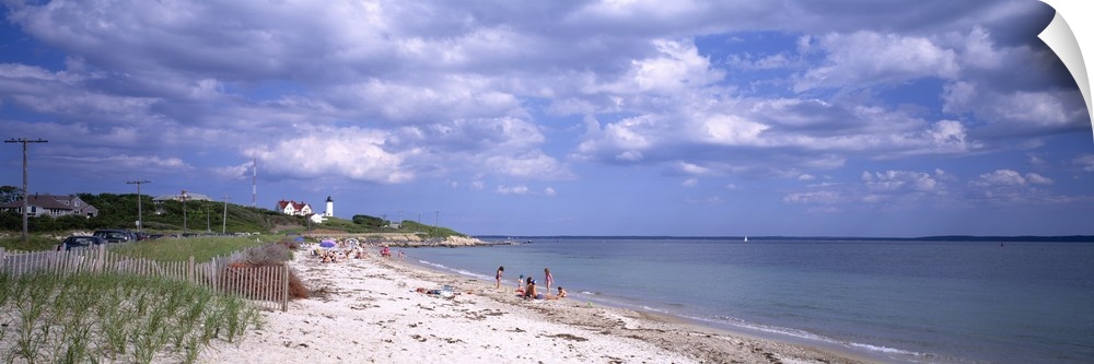 A wide angle photograph is taken of a beach in Cape Cod that only has a few people on it. A light house can be seen in the...