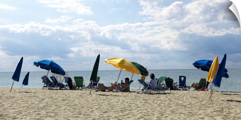Slanted beach umbrellas shade people that sit on the beach and look out over the ocean.