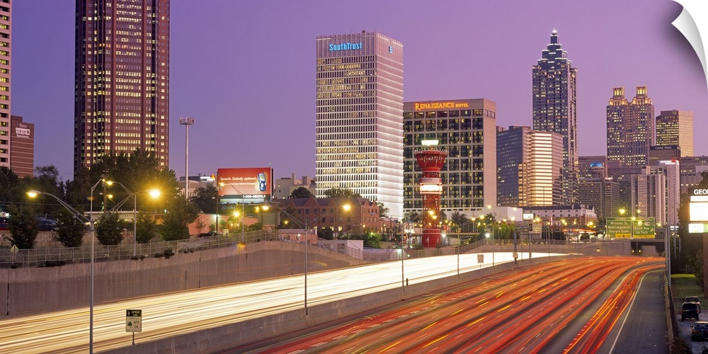 Panoramic photograph of highway filled with light trails lined by lit up buildings at sunset.