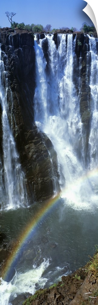 A vertical panoramic piece of an immense waterfall with a rainbow stretching across the bottom of the picture.
