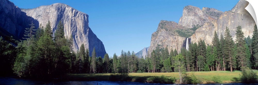 Panoramic photo of large rock cliffs and mountings surrounded by pine trees and water in Yosemite National Park.