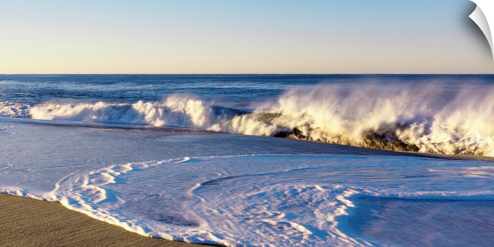 This landscape photograph shows waves curling and crashing into a sandy beach in the late afternoon.