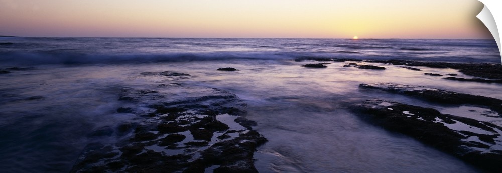 Panoramic photograph taken of the ocean as the water flows over rocks and the sun is shown setting on the horizon.
