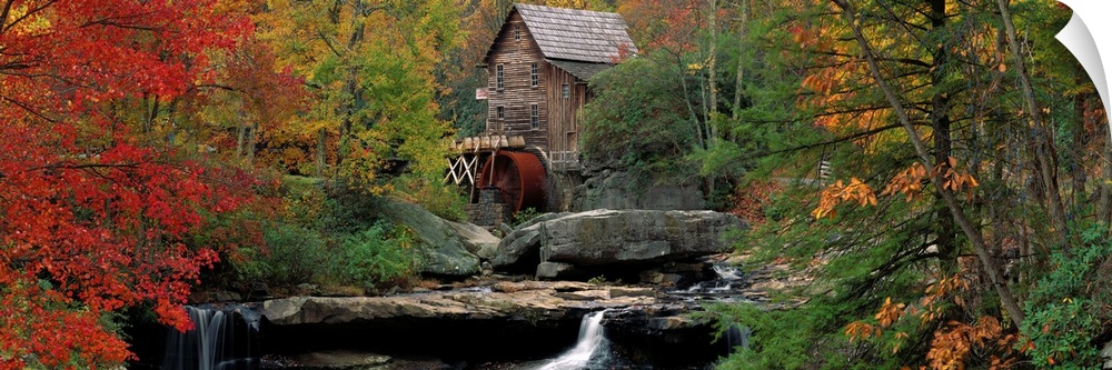 An old water mill sits at the top of flat boulders and a water fall in the center of this panoramic canvas depicting an au...