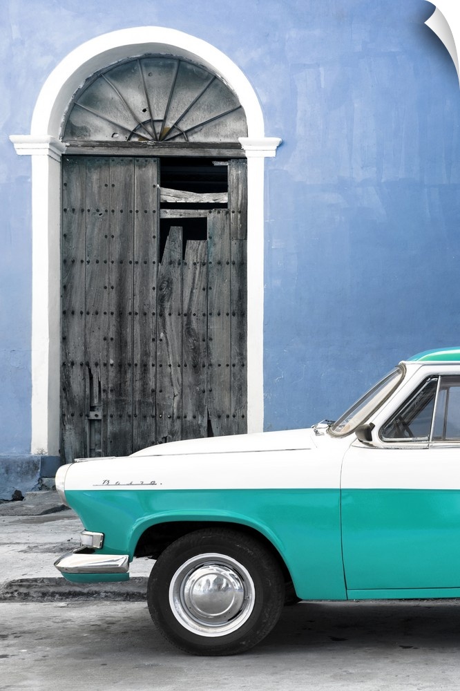 Photograph of the front of a vintage turquoise and white car outside of a blue building with a broken wooden door.