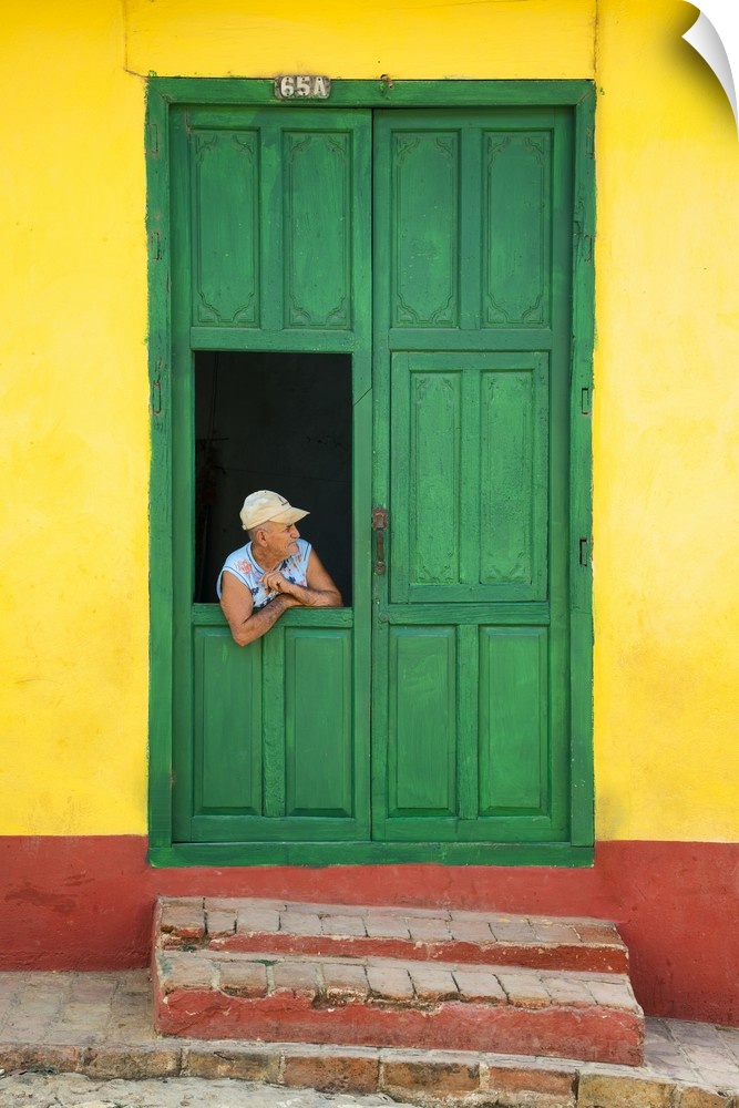 Photograph of a man leaning through a window connected to a large green door on a yellow and red wall in Cuba.