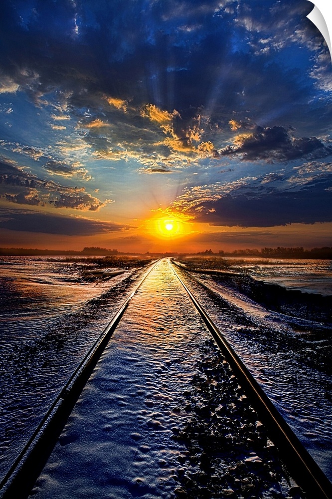 Railroad tracks leading across a snowy field to the sunset on the horizon, with a cloudy sky.