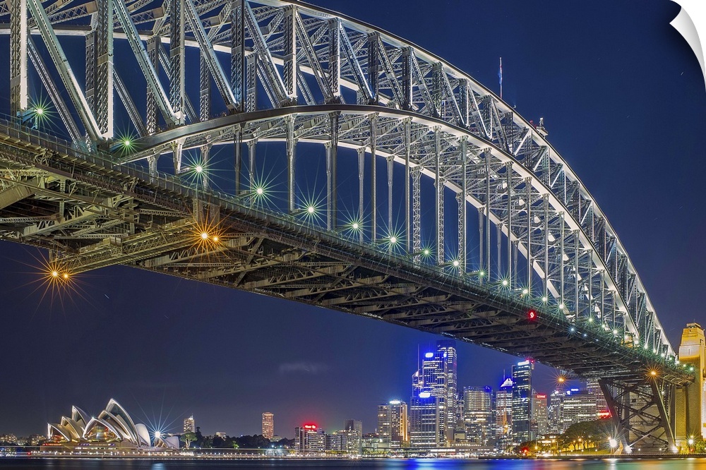 The Sydney harbor skyline lit up in neon lights at night, seen from under a large arcing bridge.