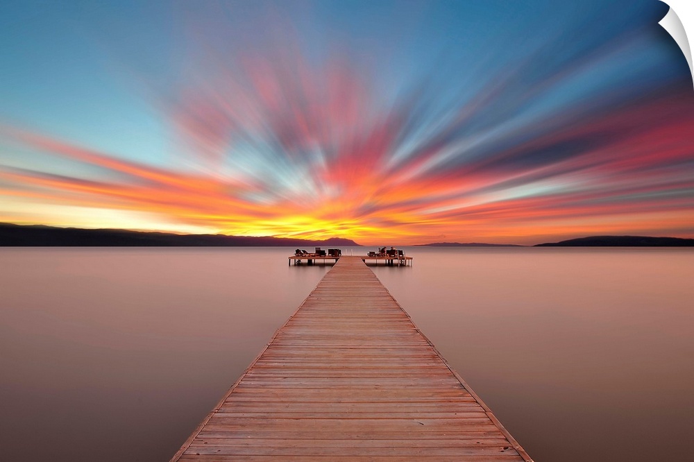 A wooden pier stretching out onto the water under a brightly colored sunrise sky.
