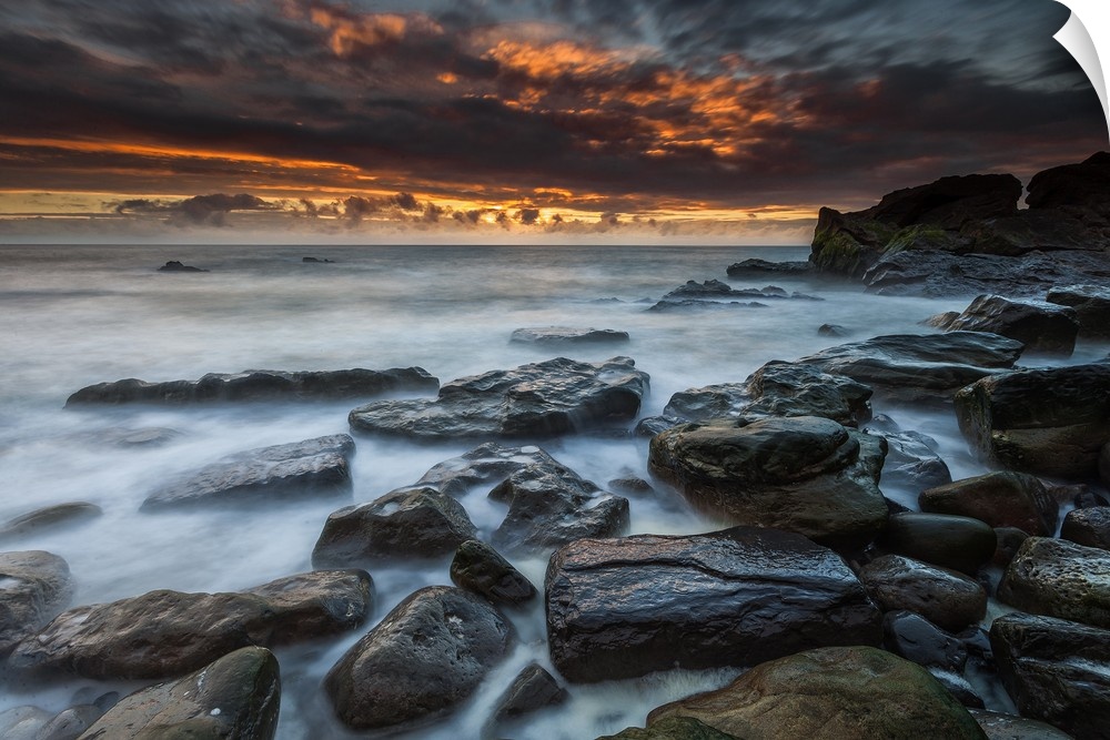 Dramatic sky filled with dark clouds illuminated by the setting sun, with a rocky coastline in the foreground.
