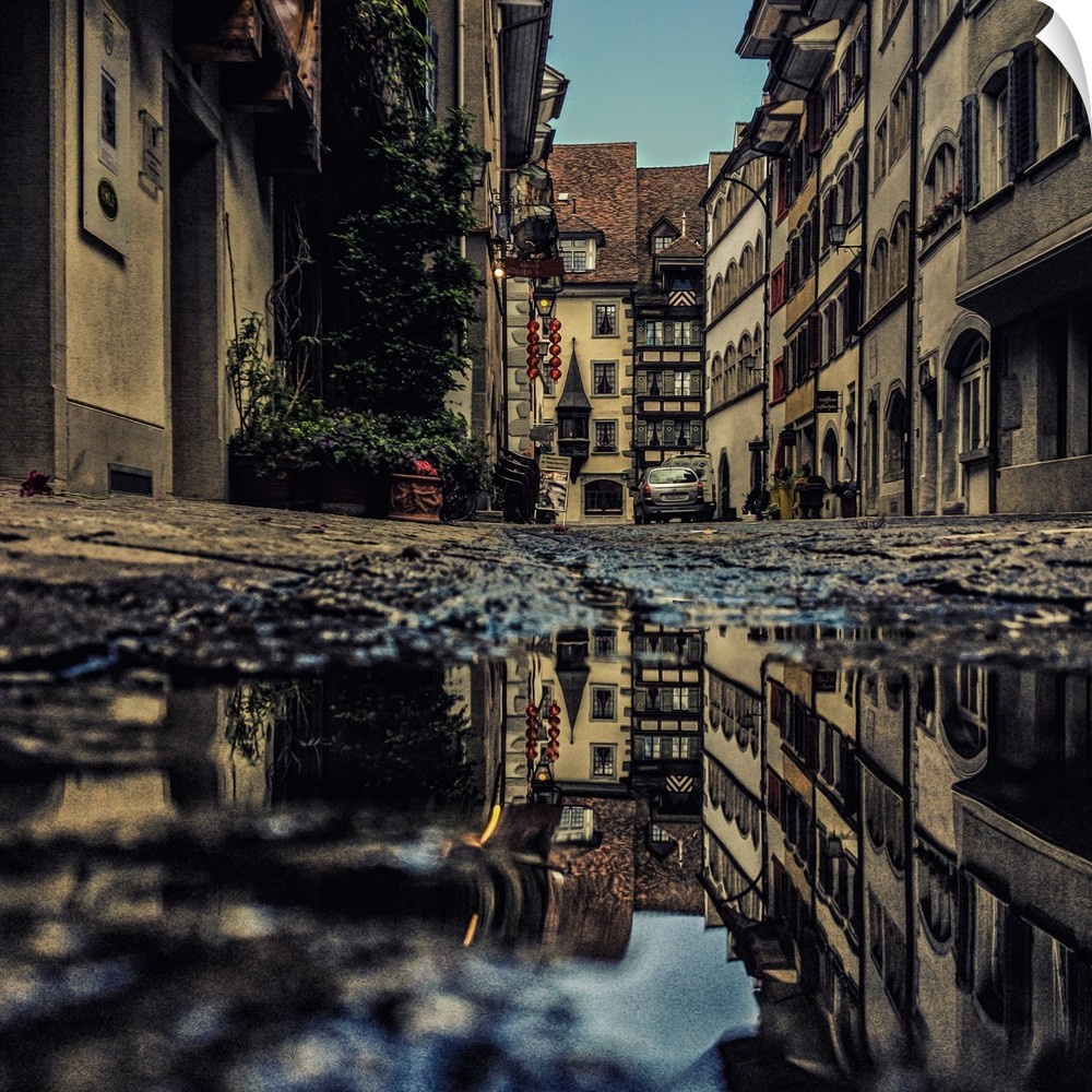 Photograph of a puddle reflecting surrounding buildings and atmosphere.