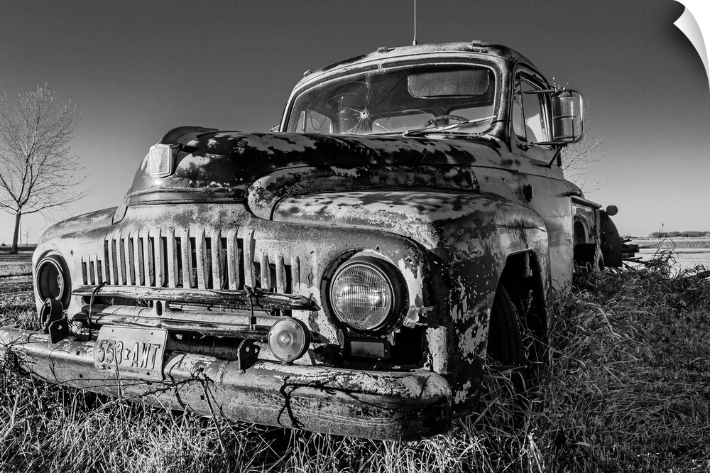 A black and white image of an old pick-up truck slowly sinking in the grass of an old farm yard at sunset.