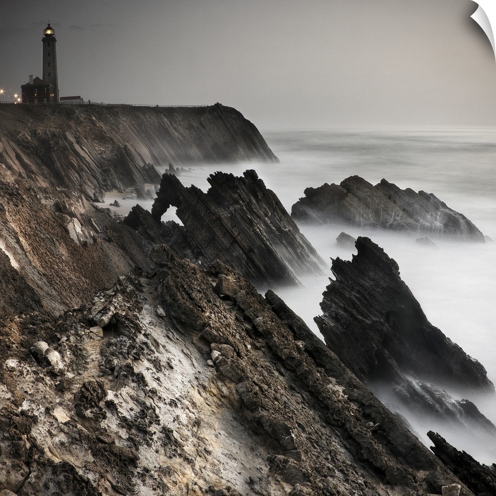 Dynamic photograph of a lighthouse on a foggy jagged rocky coast.
