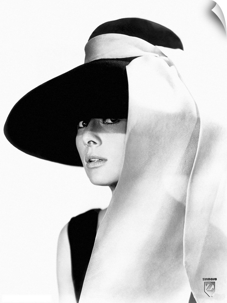 Black and white photo of Audrey Hepburn in a black dress and large floppy black hat with a white sash tied to the hat stre...