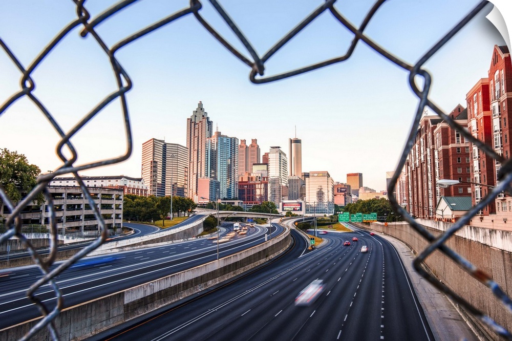 The skyline of Atlanta, Georgia, seen through a hole in a chain-link fence over a busy street.