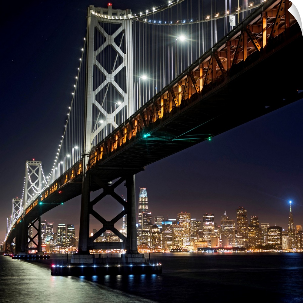 Square photograph of the Bay Bridge lit up at night with the San Francisco skyline in the background.