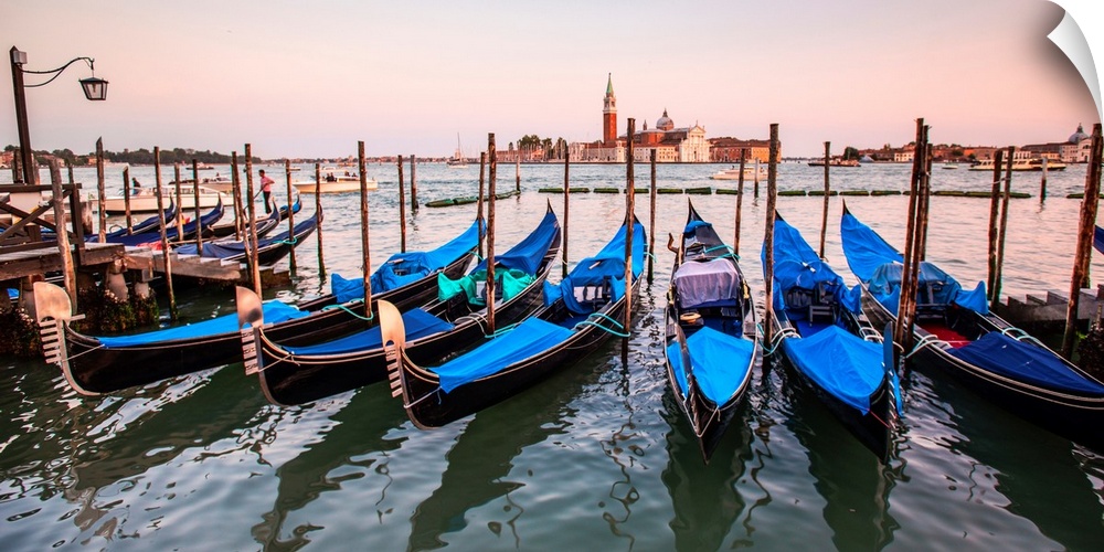 Panoramic photograph of blue gondolas docked in a row on the water with St. Mark's Square in the background at sunset.