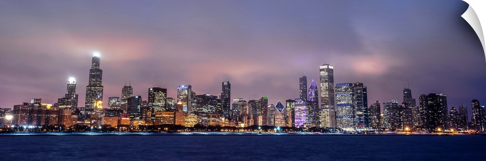 Panoramic view of the Chicago city skyline in the early evening, with city lights reflecting off the clouds above.