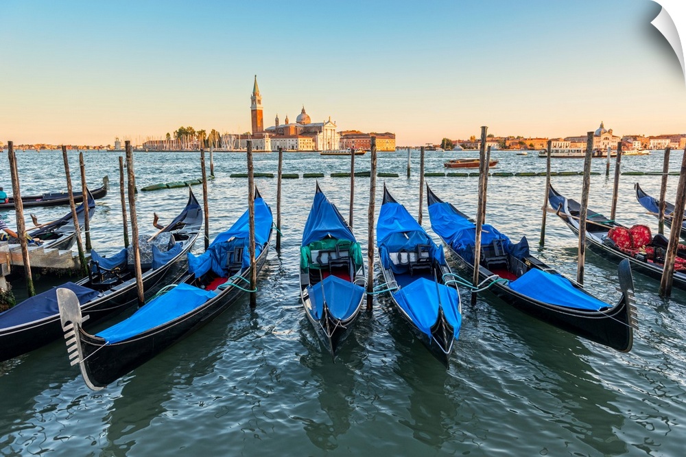 Photograph of a row of docked gondolas with St. Mark's Square (Piazza San Marco) in the background at golden hour.
