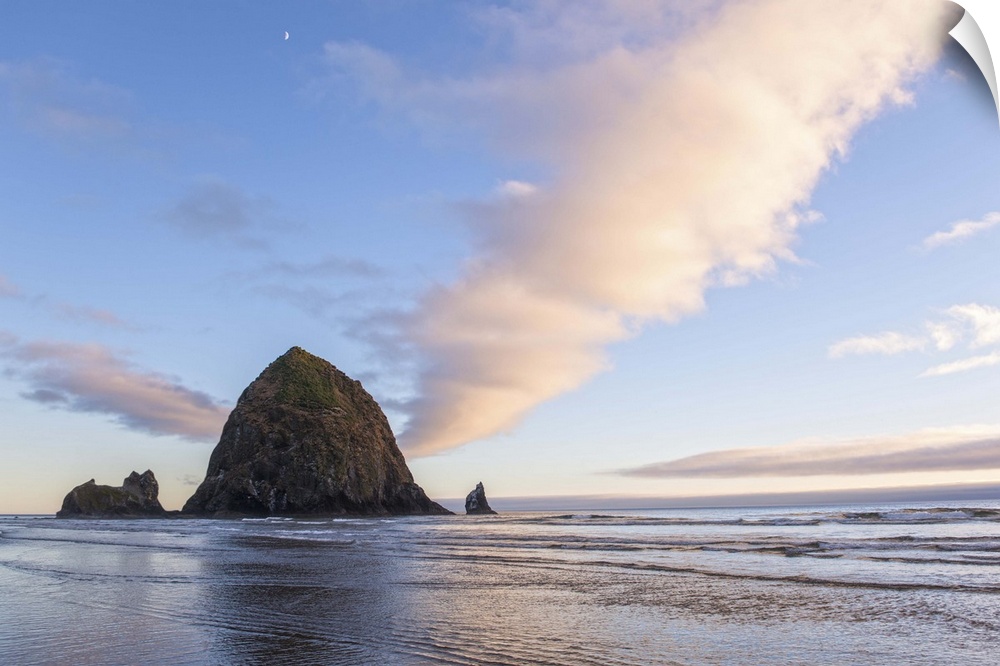 Photograph of Haystack Rock at sunset with rippling waters in the foreground and the moon above.