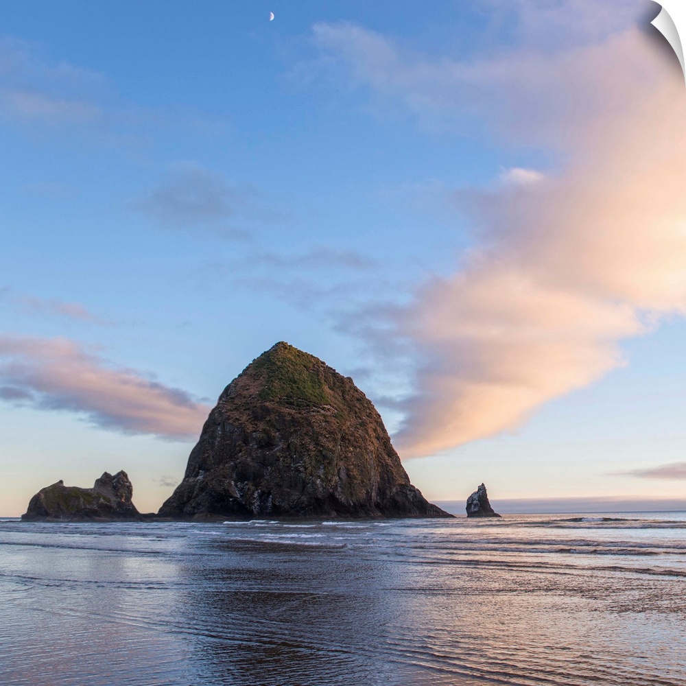 Square photograph of Haystack Rock at sunset with rippling waters in the foreground and the moon above.