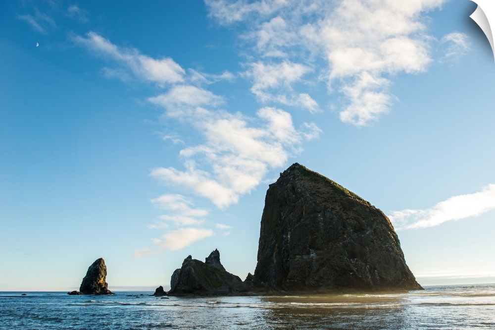 Photograph of Haystack Rock with the golden sun shining on its side and the moon in the top corner.