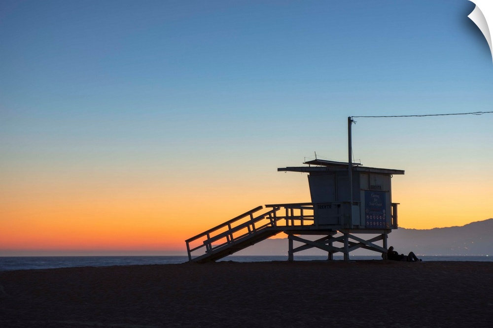 The sun sets on Venice beach with the silhouette of a lifeguard tower.