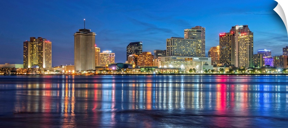 Photograph of the New Orleans skyline lit up at dusk and reflecting colorful bands onto the Mississippi River.