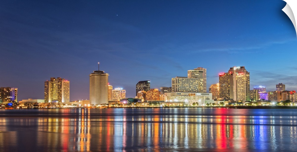 Photograph of the New Orleans skyline lit up at night and reflecting colorful bands onto the Mississippi River.